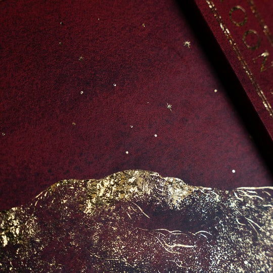 A close-up of the 13 moon journal 2027 dark burgundy textured surface with scattered small metallic gold star-like specks and a chunky gold foil mountain-like border along the bottom edge.