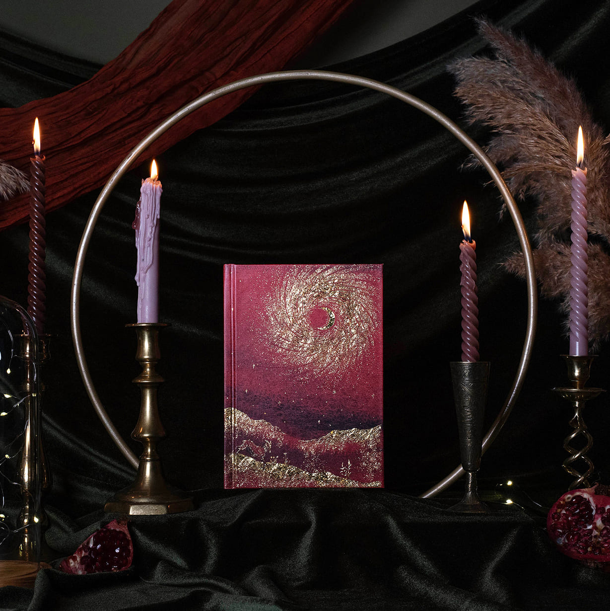 A s13 moon journal 2027 cover featuring a swirling galaxy design in deep magenta and gold, styled on a black fabric backdrop with lit candles, a metal hoop, and pampas grass.