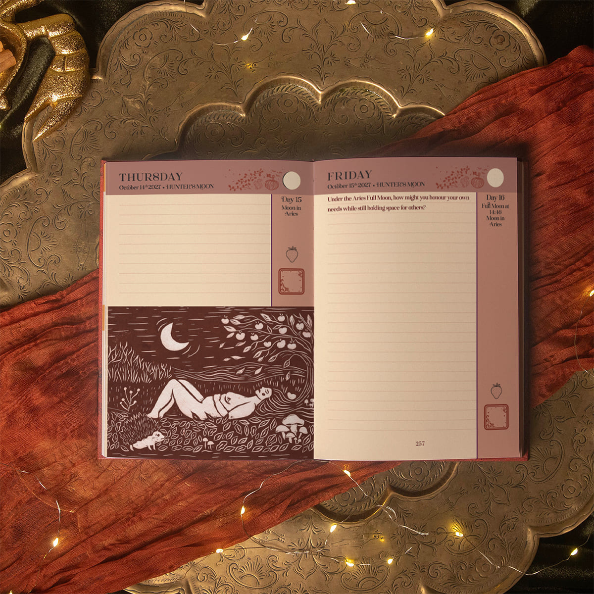 An open weekly planner spread with lined Thursday and Friday pages, a dark brown illustration of a woman lying under a crescent moon, and a dusty mauve header, styled on a carved brass tray with rust-colored fabric and warm string lights.The 13 Moon jOurnal 2027 