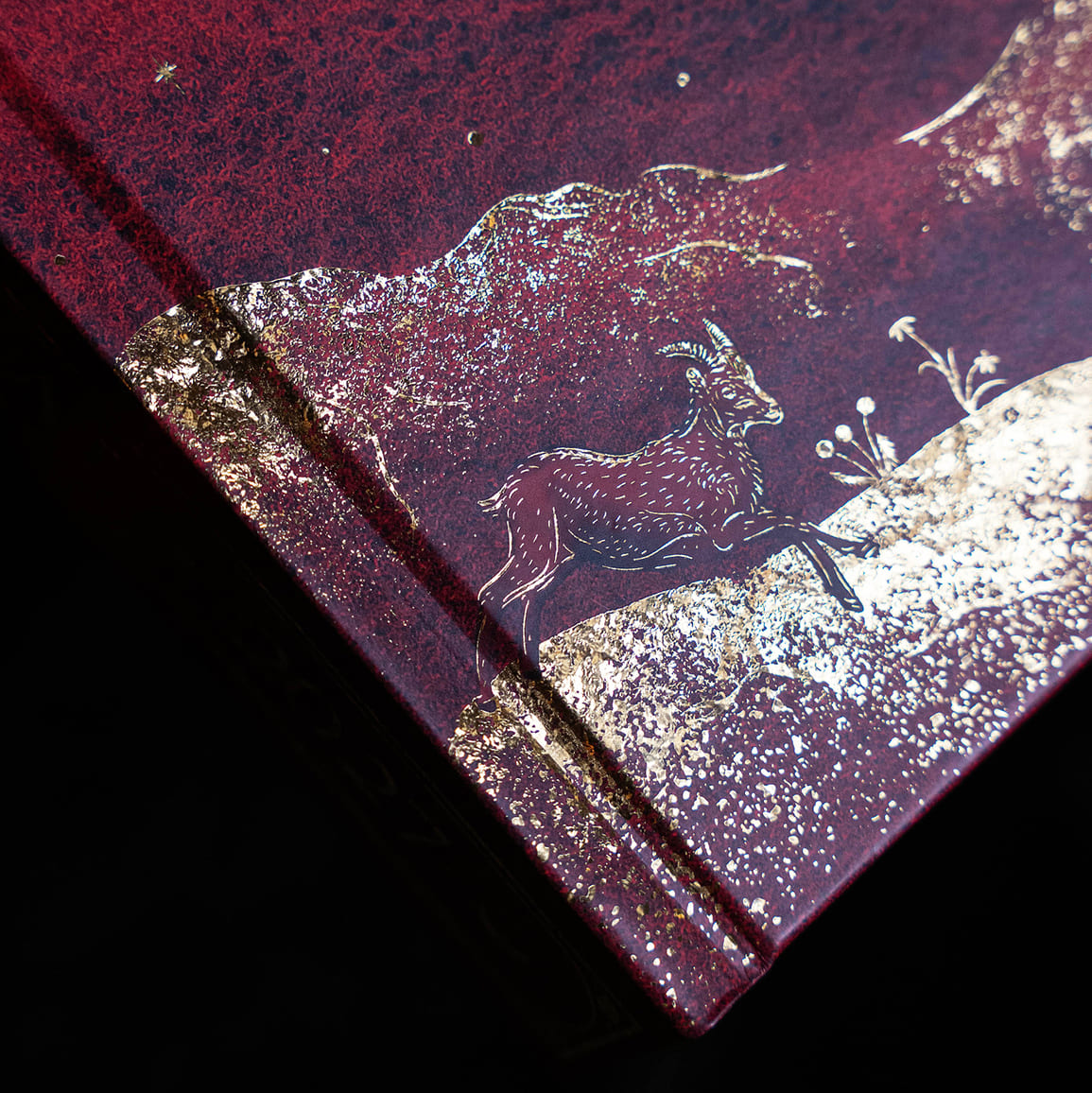 A close-up of a textured dark red or burgundy surface with metallic gold foil detailing, featuring an embossed deer and small floral motif against a black background. Moon Journal 2027