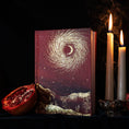 Load image into Gallery viewer, A hardcover book with a mystical cover illustration of a swirling golden vortex in a starry burgundy sky, styled on a dark cloth next to a halved pomegranate and two lit white candles.
