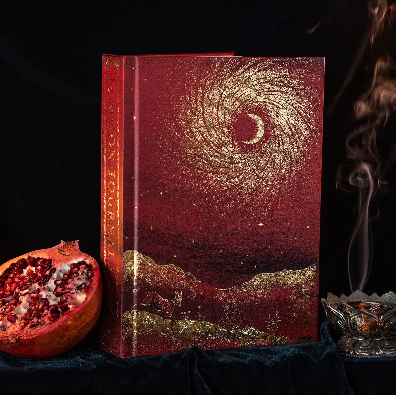 A decorative hardcover 13 moon journal 2027 with a deep red and gold celestial cover design featuring a crescent moon and swirling star pattern, displayed on a dark velvet surface next to a sliced pomegranate and a smoking incense burner.