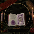 Load image into Gallery viewer, An open 13 moon journal 2027 book displayed on a stand, with the pages showing purple and white card art and text. The book is styled in a dark, mystical setting with a gold hoop, lit candles, green velvet fabric, and dried pampas grass.
