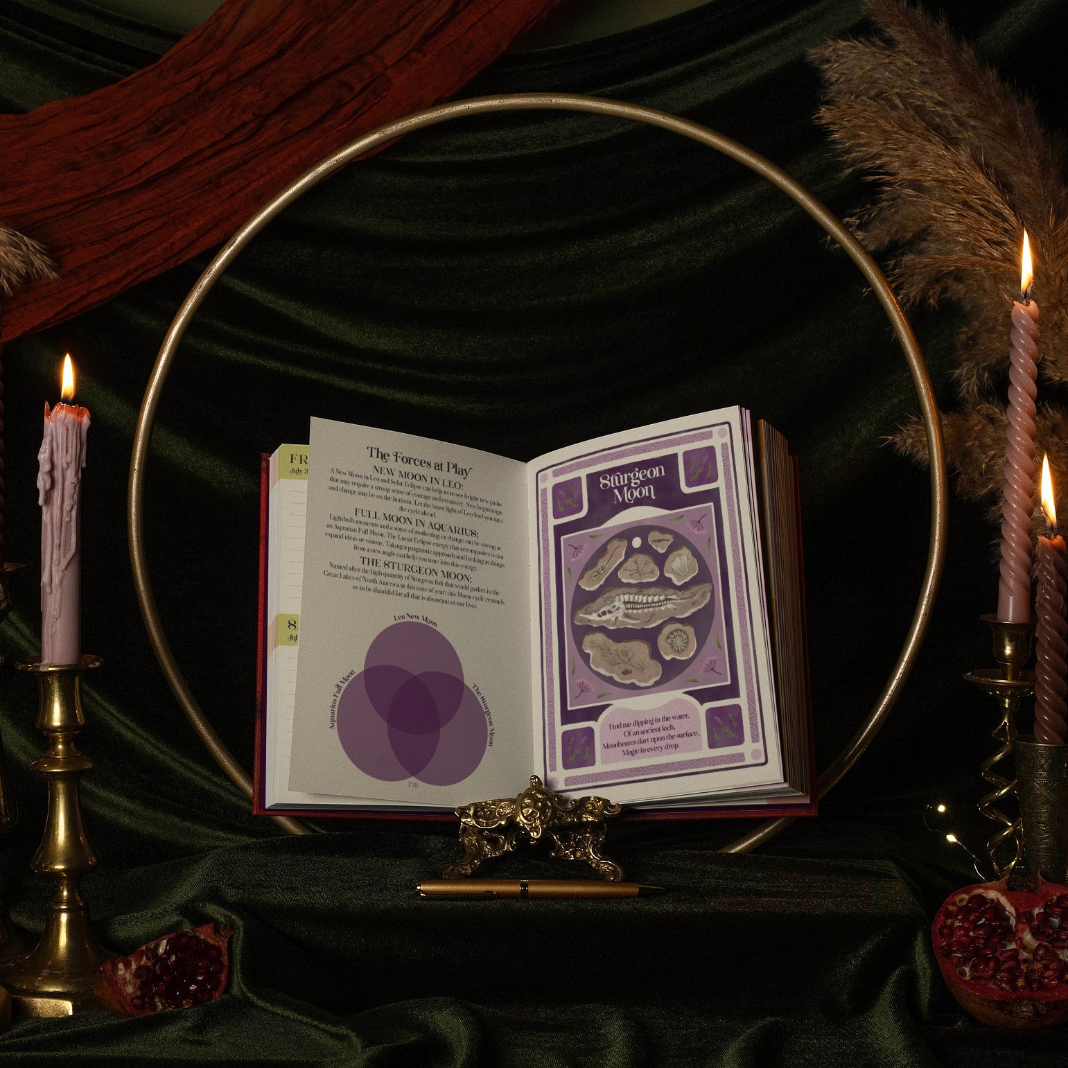 An open 13 moon journal 2027 book displayed on a stand, with the pages showing purple and white card art and text. The book is styled in a dark, mystical setting with a gold hoop, lit candles, green velvet fabric, and dried pampas grass.