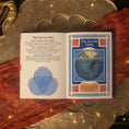 Load image into Gallery viewer, An open book spread laid on a metallic tray with draped red fabric and warm string lights. The left page has text titled 'The Forces at Play' with a blue Venn diagram, and the right page has a titled illustration panel reading 'The Beaver Moon.
